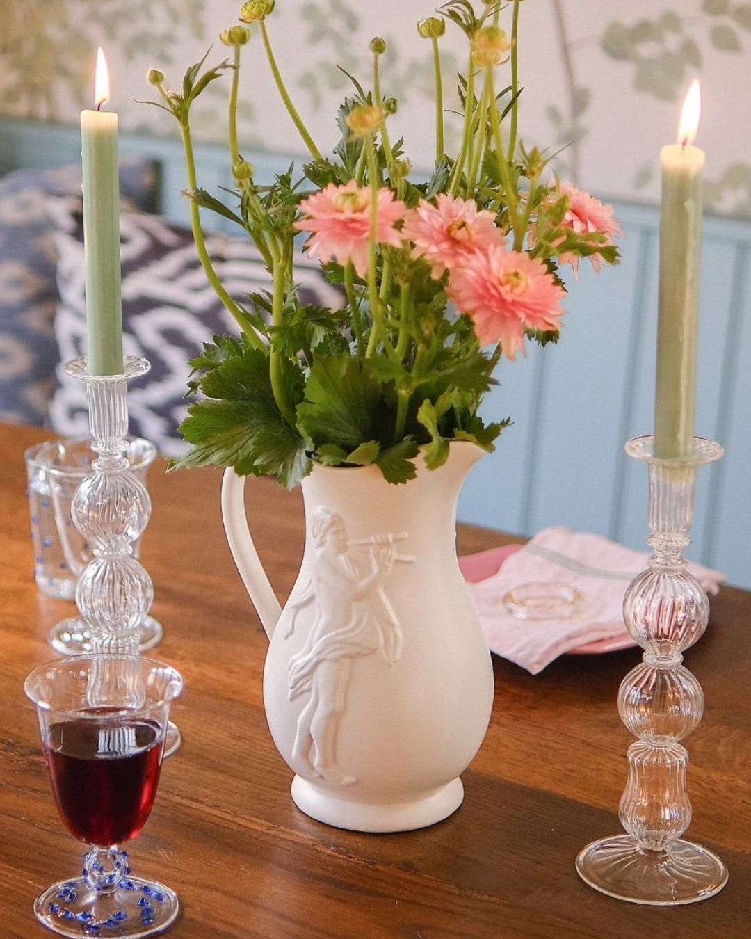 White pitcher with flowers, two lit candles, and a glass of red wine on a wooden table. Issy Granger
